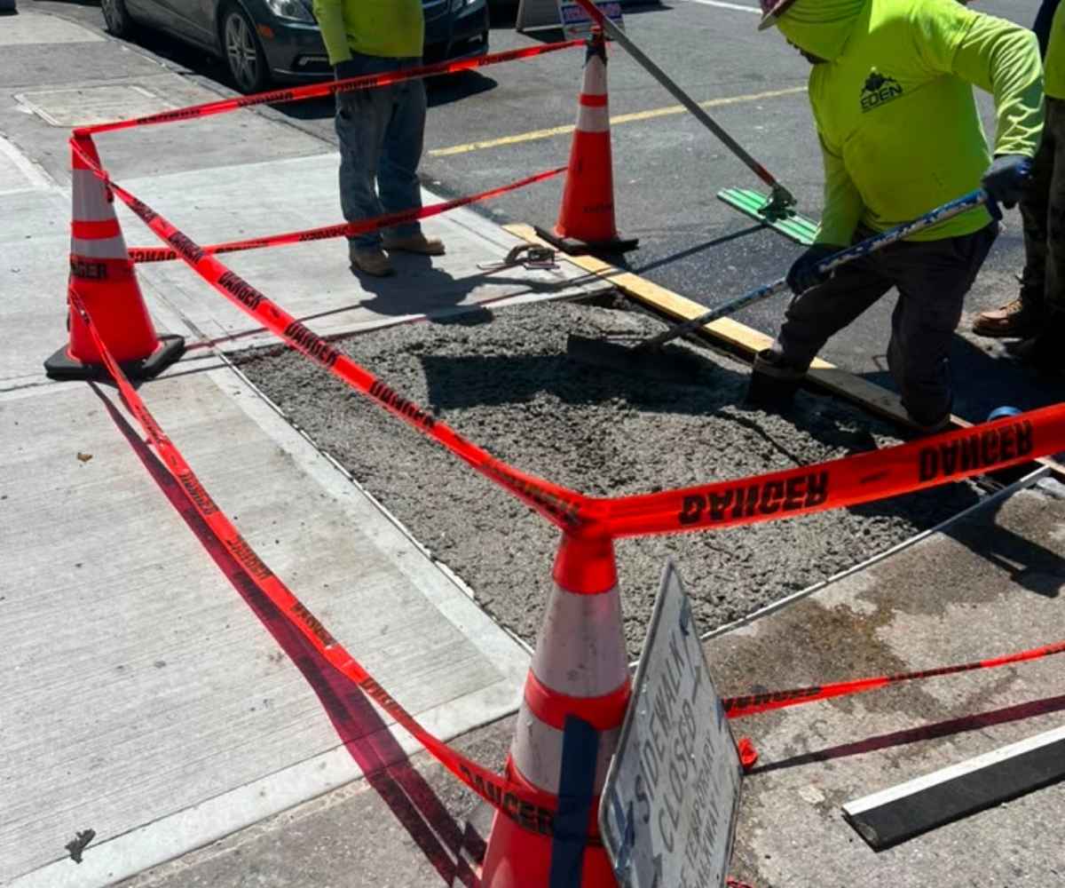 Crew finishing a fresh concrete sidewalk pour inside a taped-off work zone in Brooklyn